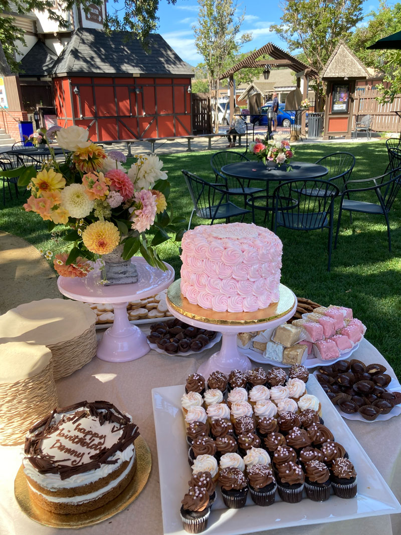Birthday cakes and cupcakes with other desserts on a table with park in the background