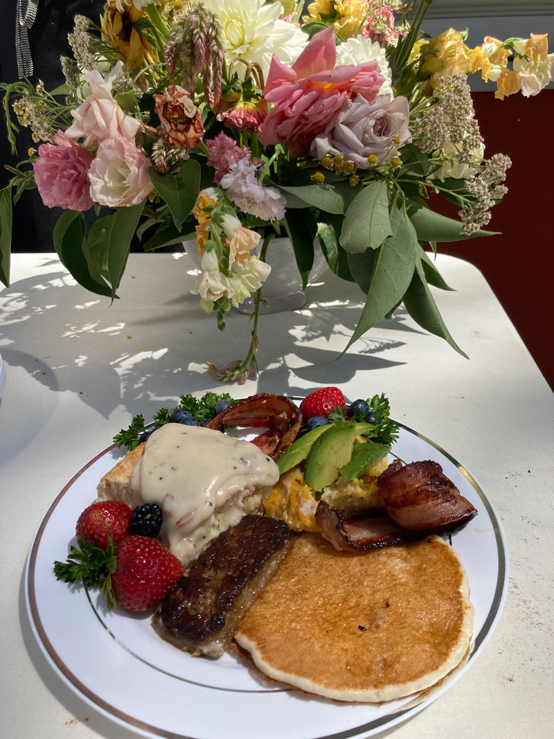 floral bouquet behind a plate with pancake, fruit, sausage, bacon, and biscuit with gravey