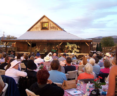 Large group of people sitting around tables at outdoor event in front of barn with musicians.